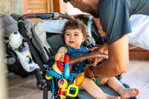 Father securing his smiling baby daughter in a grey stroller with colorful toys, readying them for