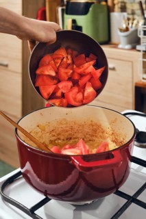 Person adds chopped tomatoes into cast iron pot on stove. Pot contains onions and spices. Kitchen