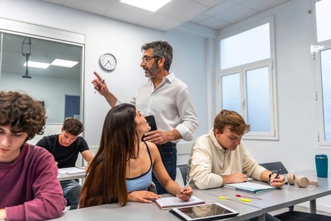 Teacher engaging female student at front of bright classroom while peers take notes and study