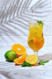 A glass with a drink made from orange and lime sits on a marble table. Fresh orange and lime slices