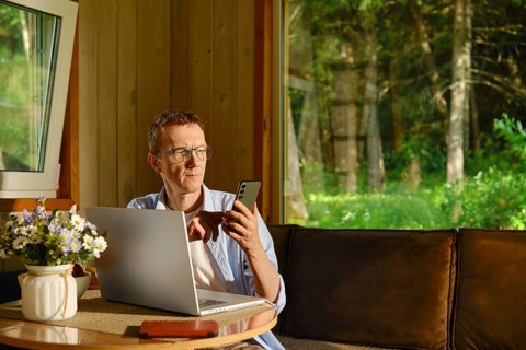 Middle-aged nomad sits at table in wooden cabin using laptop and smartphone. Sunlight shines