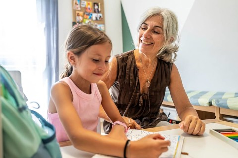 Grandmother sitting at a table, smiling and supervising her granddaughter coloring in a book,
