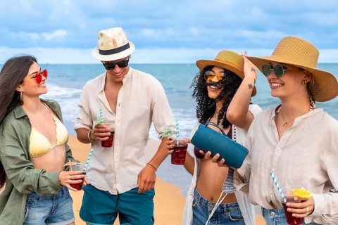 Young friends walking along a sunny sandy beach, carrying drinks and a portable speaker, laughing