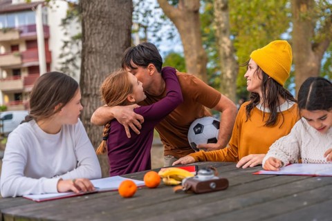Family spending time outdoors at a park picnic table, father hugging one daughter while holding a