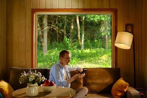 Middle-aged man sits on couch in wooden cabin, holding electronic book and reading a novel. Light