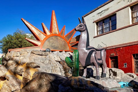 Colourful statue of a llama next to a large sun symbol and cacti on rocks in front of a building,
