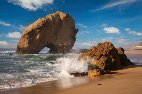 Majestic Penedo do Guincho rock arch rises above crashing ocean waves and golden sands at Praia da