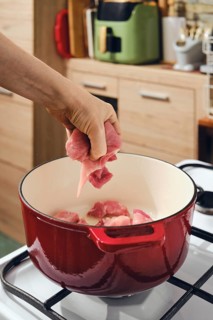 Person adds raw meat in red cast iron pot on stove in kitchen. The pot sits on gas burner in