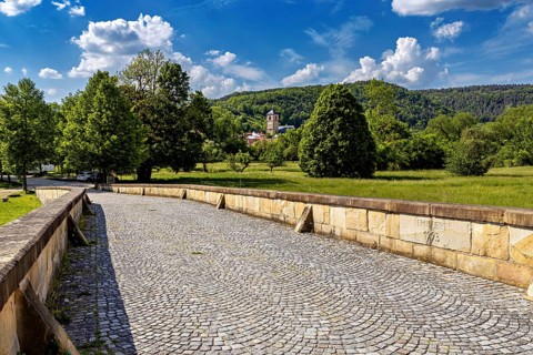 Stone bridge leads through a green landscape with trees and a village in the background, view of