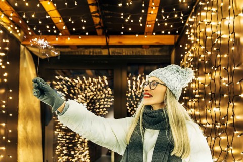 Young woman holding a sparkler with festive lights. A woman wearing a gray knit hat, dark scarf,