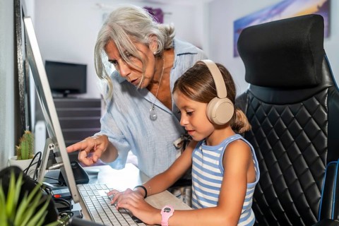 Grandmother is patiently teaching her young granddaughter how to use a computer. Pointing at the
