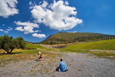 Two people sit in the courtyard of an ancient stadium with mountain background, Messene Stadium,
