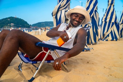 Young man relaxing on a beach chair under striped umbrellas, smiling and applying sunscreen to his