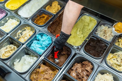 Person's gloved hand scooping chocolate gelato from a vibrant, multicolored display case, featuring