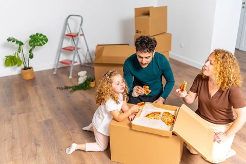 Family enjoying a pizza meal on the floor of their new, unfurnished house, surrounded by cardboard