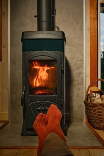 Feet in red socks rest in front of wood stove with flames. Basket nearby shows logs. Room has