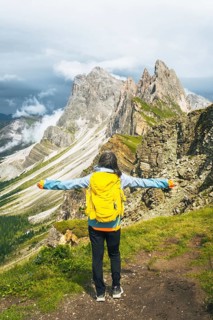 Back view middle aged gray hair woman hiker arms outstretched with yellow backpack overlooking lush