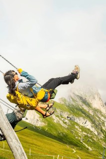 Excited woman with backpack swinging on wooden playground swing lush green hills steep mountain