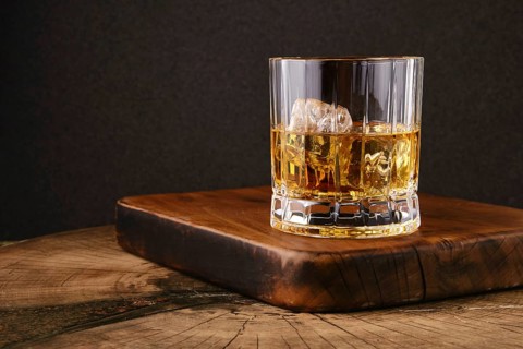 Facetted glass filled with whiskey and ice on wooden board in kitchen. Dark background highlights