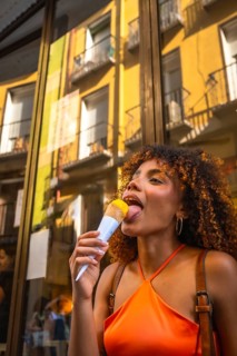 Young woman with curly hair wearing an orange top licking a yellow ice cream cone, experiencing
