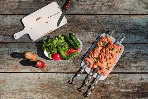 Top view of marinated chicken pieces on skewers beside tomatoes, cucumbers, and herbs on wooden