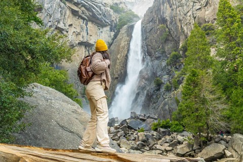 Woman standing on a fallen log, looking at the powerful flow of lower yosemite falls and feeling
