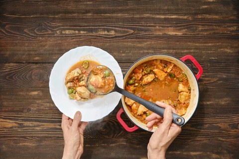 Top view of person using ladle to serve meat stew into white plate. Pot is full of stew with