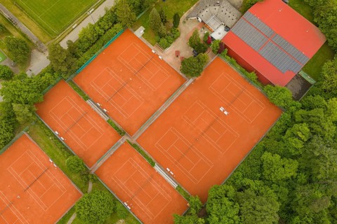 Tennis courts with orange sand in an organized sports complex, surrounded by buildings and nature,