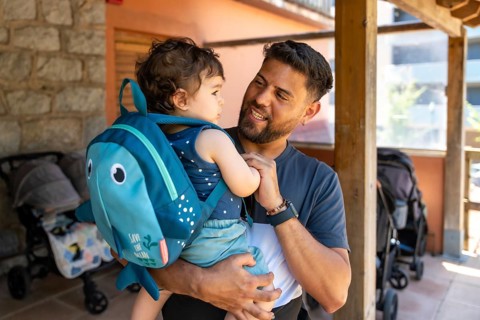Father holding his young son, who is wearing a fish shaped backpack, smiling and looking at him