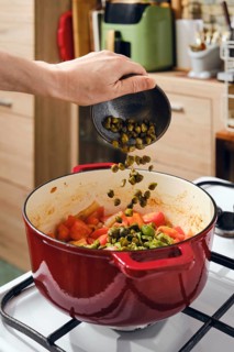 Hand pours capers into pot filled with chopped tomatoes, onion and green olives. Pot sits on stove