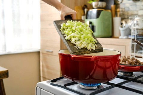 A hand holds a cutting board with chopped celery above a red pot on a stove. The kitchen has