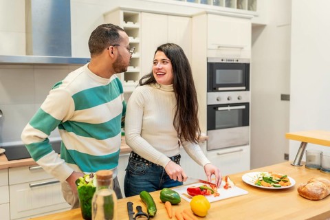 Young happy couple standing in a bright kitchen, the woman preparing fresh vegetables while looking