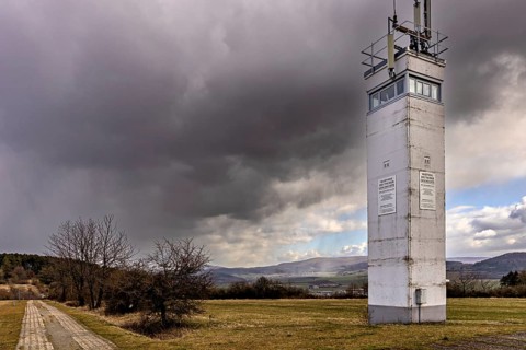 A watchtower stands under a dramatic cloudy sky in a meadow next to a small road, The Point Alpha