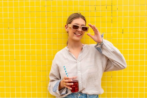 Young woman leaning on a vibrant yellow tiled wall, adjusting her sunglasses with one hand while