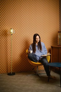 Woman with long hair sits in yellow chair. She holds book in one hand and cup in other. Room has