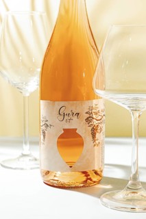 A bottle of Gura wine is placed on a table beside two empty wine glasses. Natural light shines