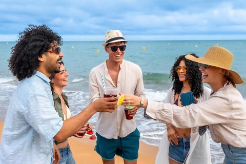 Group of diverse young adult friends cheering with colorful drinks on a sandy beach, enjoying