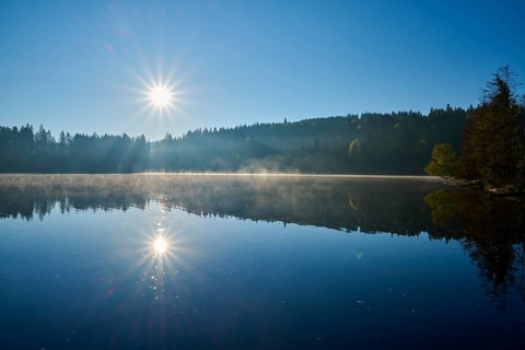 The sun rises over a still lake with light fog on the water surface, Windgfällweiher, Lenzkirch,