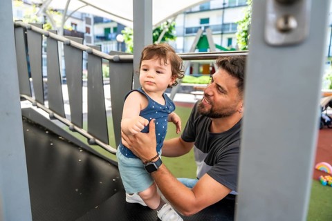 Father guiding toddler on playground structure during sunny park playtime, sharing joyful bonding,