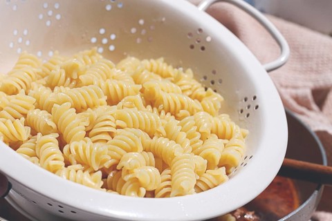 A person is holding a colander filled with cooked rotini pasta. Water is draining off as the pasta