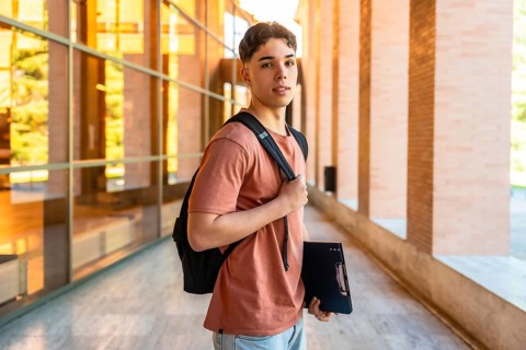 Male student walking along modern campus building with backpack and folder, reflecting college life