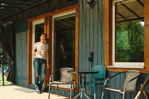 Relaxed middle-aged woman stands on the porch of wooden cabin holding drink. There are two chairs