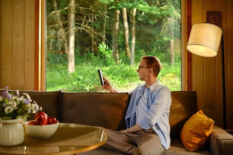 Middle-aged man sits on brown sofa in wooden cabin. He reads e-book in air-conditioned room while