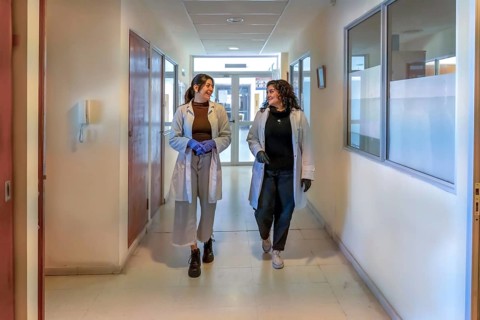 Two young female scientists in white lab coats and gloves smiling and talking while walking