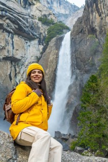 Woman in a yellow jacket and beanie with a backpack sitting on a rock, smiling at the camera,