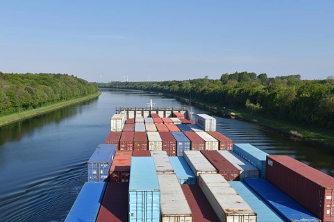 Container ship sails through green river, surrounded by forest, under clear skies, container ship