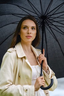 Woman stands outside with black umbrella in hand. She is wearing a light coat and looks thoughtful
