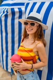 Young woman wearing a hat and sunglasses, smiling while enjoying a cold beverage on a sunny summer