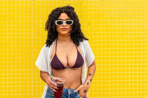 Young woman wearing bikini top, white cover up, and sunglasses, standing against a vibrant yellow