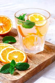 Slices of orange and lemon are arranged on a wooden board next to a glass of drink. Fresh mint